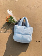 Mini Puffer Tote Bag in light blue with gingham strap on sandy beach beside orange flowers