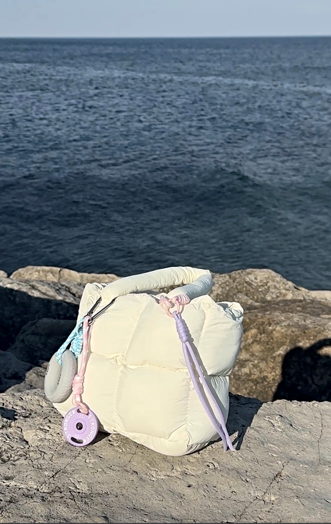 Mini Puffer Tote Bag in cream with pastel purple and blue straps and charm, on rocky shoreline