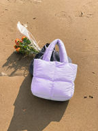 Mini Puffer Tote Bag in lilac purple on sandy beach beside orange flowers